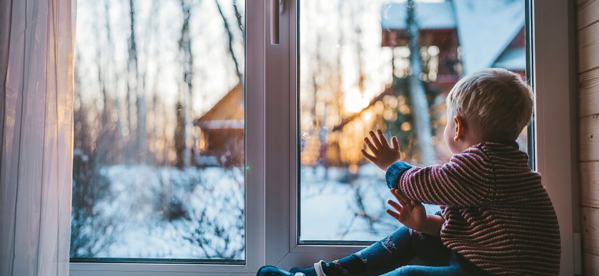 little boy looks out the window at home in winter waving his hand and feeling sad on a cold day