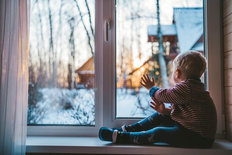 little boy looks out the window at home in winter waving his hand and feeling sad on a cold day